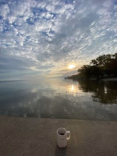 White coffee mug sitting on a lakeside ledge at sunrise, with the sky full of soft clouds reflecting on the still water, creating a peaceful and serene scene.